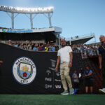 Técnico Pep Guardiola entrando em campo -há do lado esquerdo uma bandeira do Manchester City com fundo preto no lado esquerdo da imagem - o City acertou a contratação do Marc Guéhi nesta sexta-feira, 16/01/2026